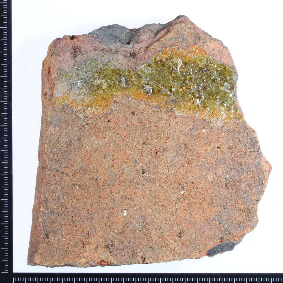 Et rødbrændt fragment af en gulvflise med rester af grøn/gullig glasur på oversiden. Mål: 9x9x2 cm.