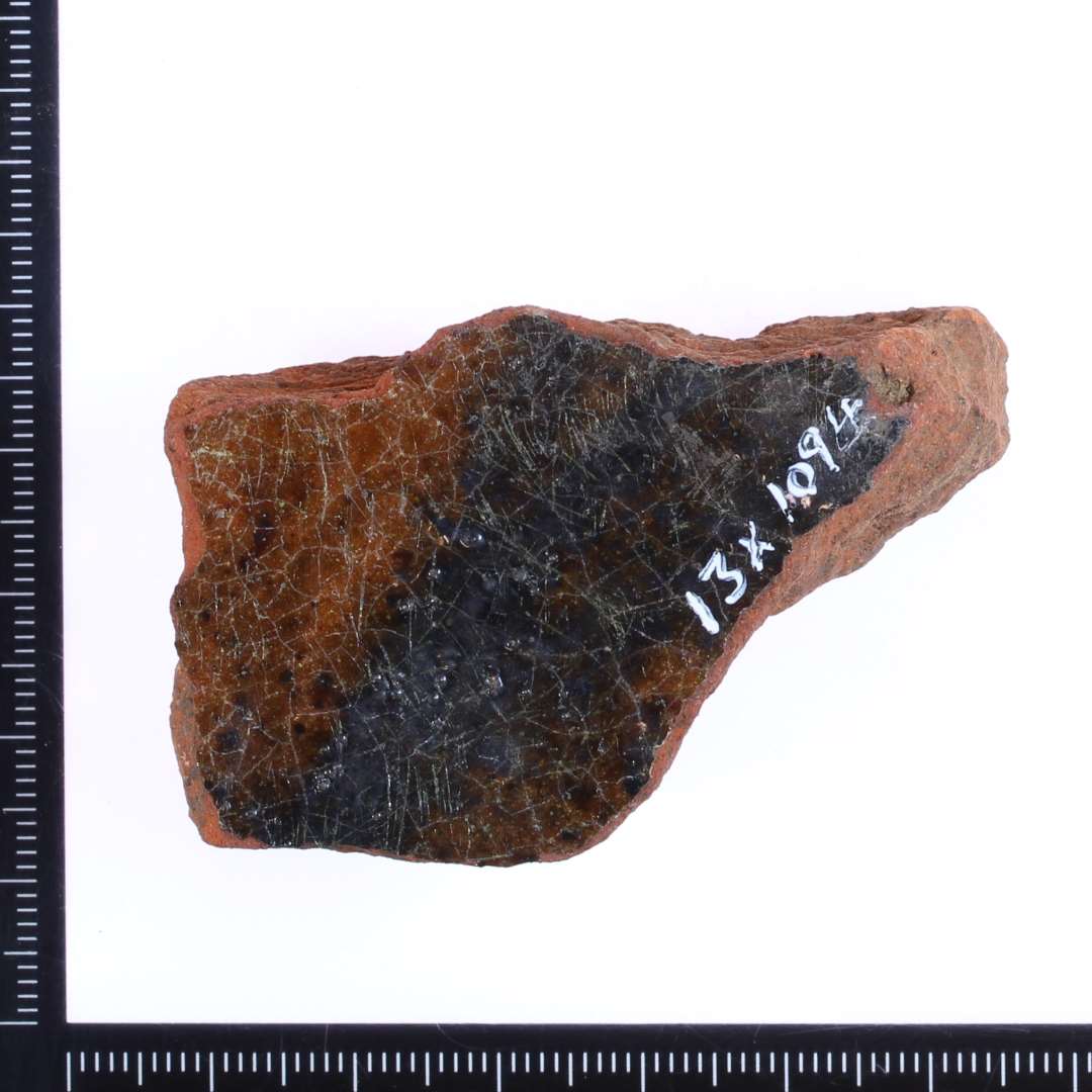 Et lille rødbrændt fragment af en gulvflise med grøn/rødlig glasur på overside. Mål: 5x3,5x2 cm.