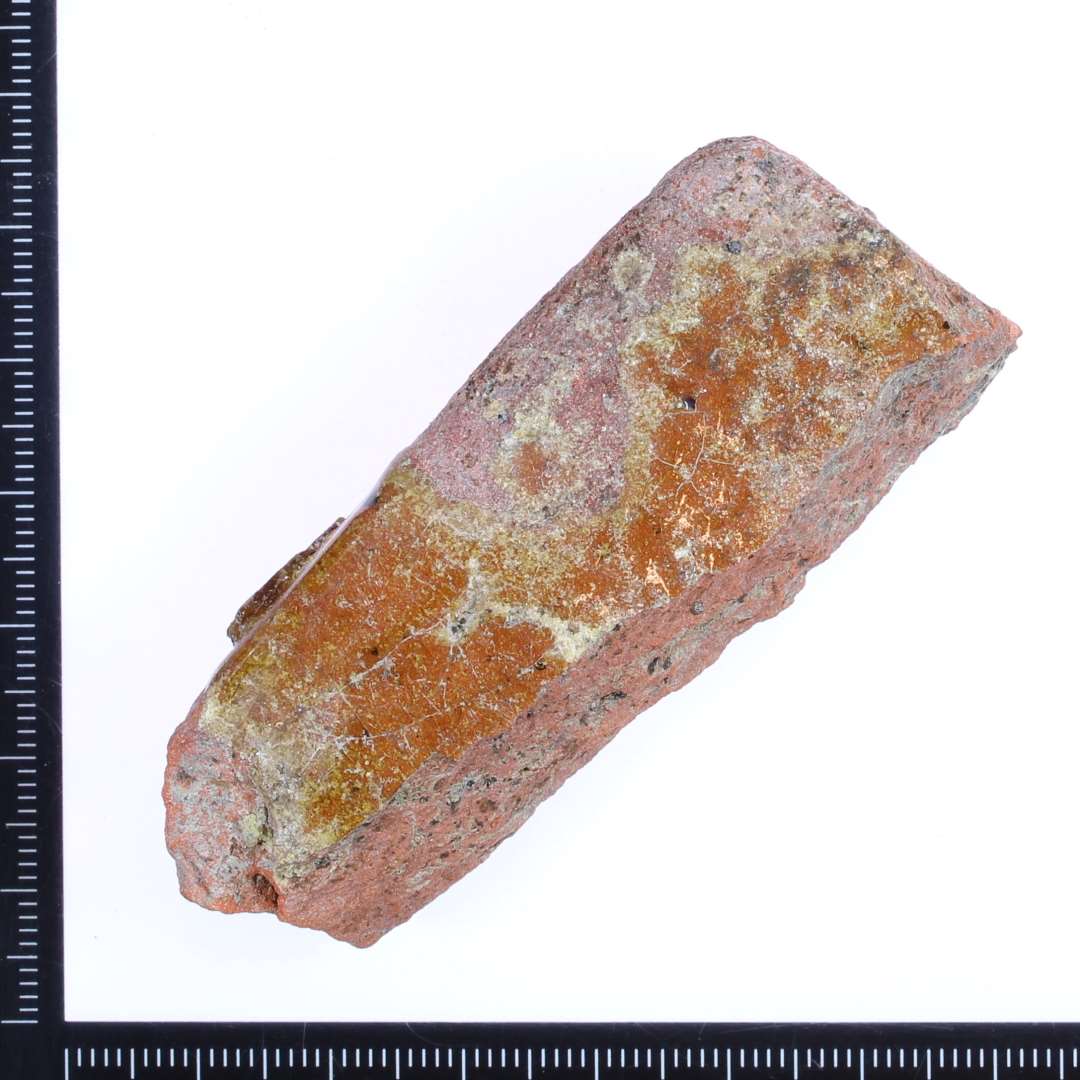 Et lille rødbrændt hjørne fragnment af en gulvflise med rester af rød/gullig glasur på oversiden. Mål: 7x2,5x2 cm. 