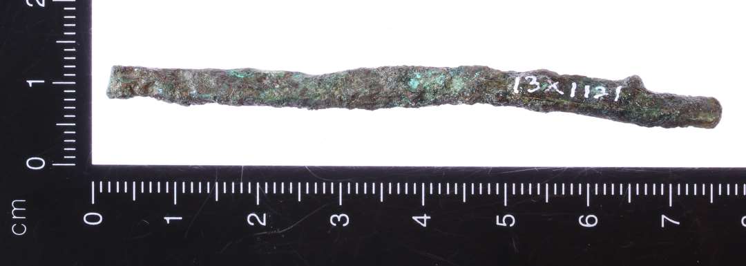 7,5 cm lang 0,5 cm bred bronze/kobber stykke. Indsnævning i den ene ende, samt en mindre indsnævning i den anden ene, så denne får en 'kødbensform'. Muligvis dorn til spænde e.lign.