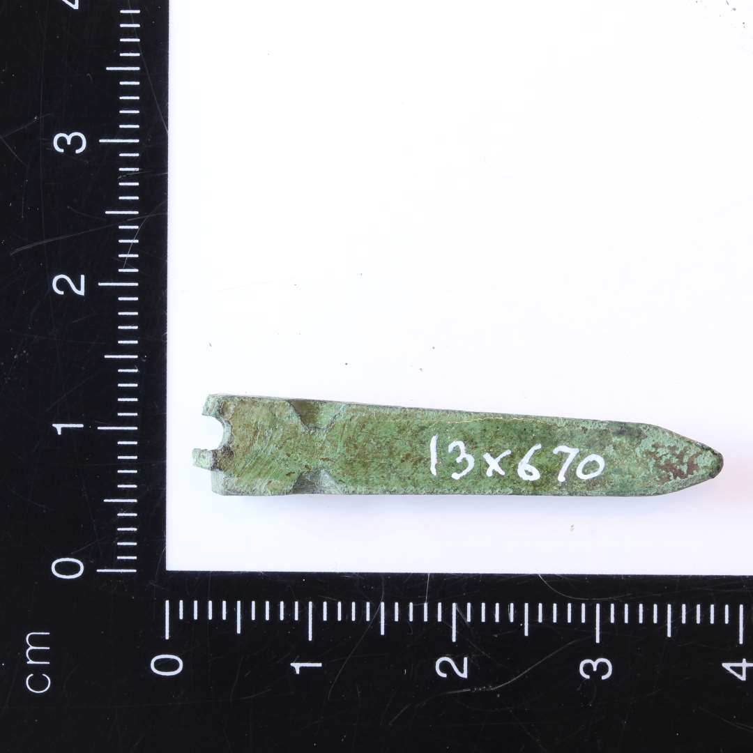 3,5 cm langs tilspidset bronzegenstand. Fragmentet i den ene ende, men med rester af hul i samme ende. Affasede kanter på spidsen. Formodentlig kraftig bæltedorn.