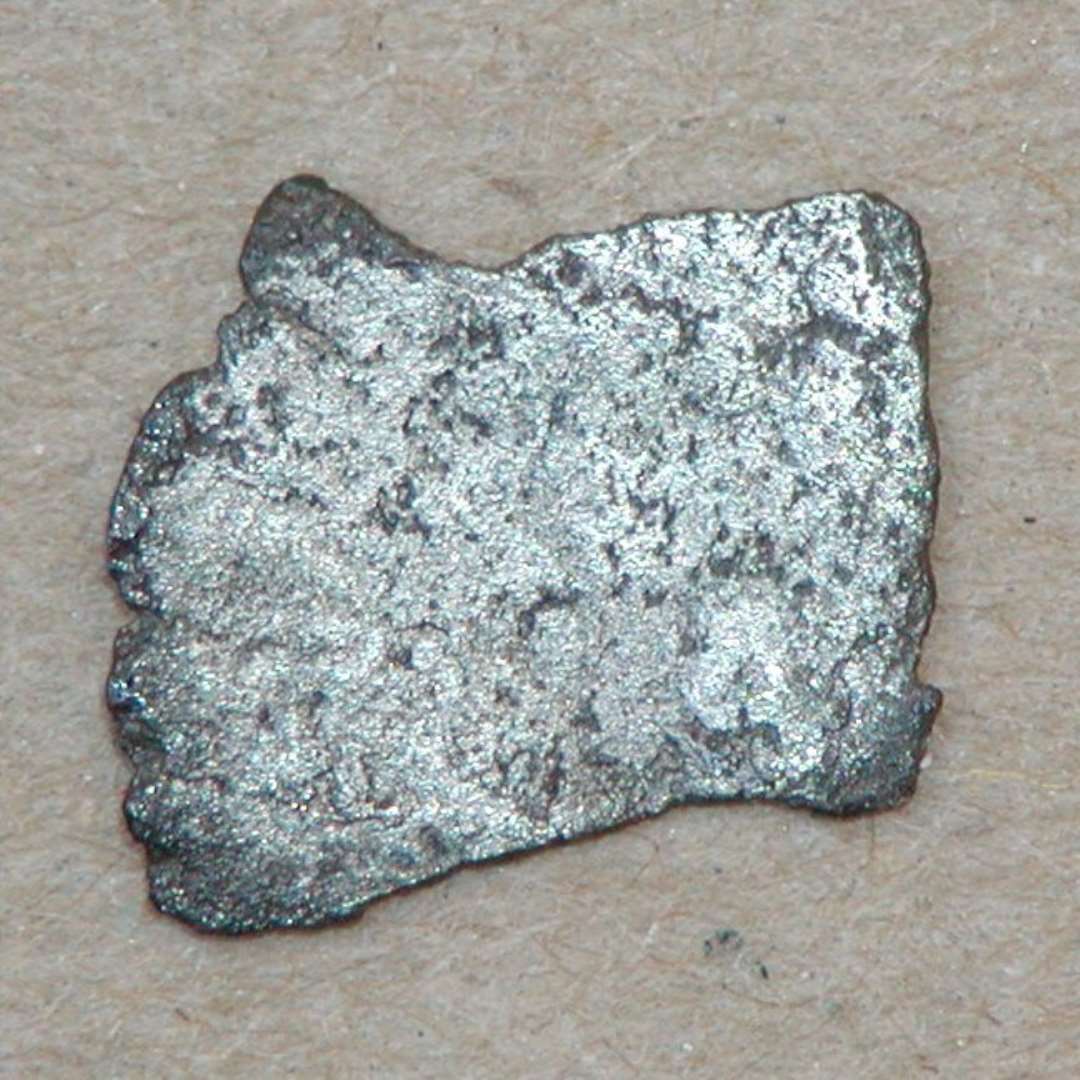 Mindre, meget slidt fragment af formodet kufisk mønt, ca. 11x8 mm. På den ene side ses svage skrift-tegn. Dirhem.