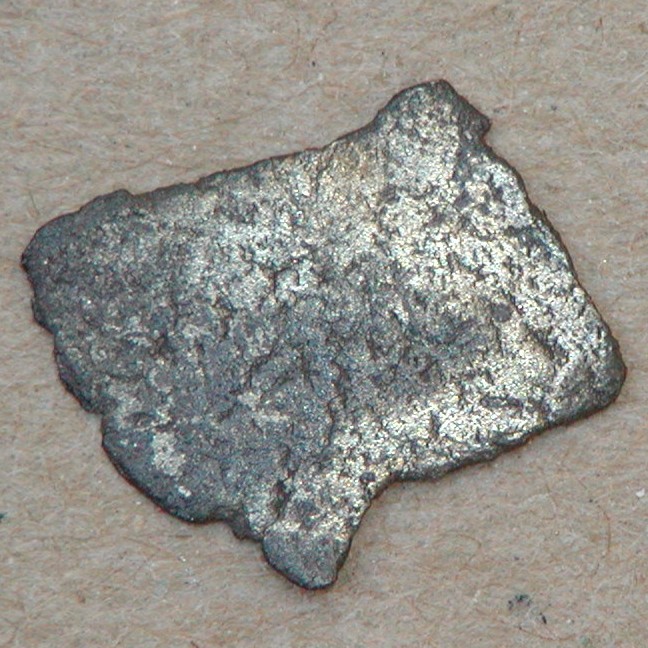 Mindre, meget slidt fragment af formodet kufisk mønt, ca. 11x8 mm. På den ene side ses svage skrift-tegn. Dirhem.