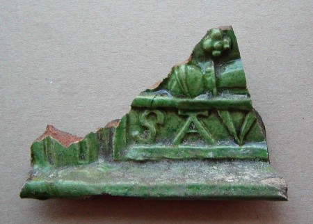 Fragment af grønglaseret kakkel. Nedre del med liste og felt med bogstaverne 'SAV'. Ca. 11x7 cm.