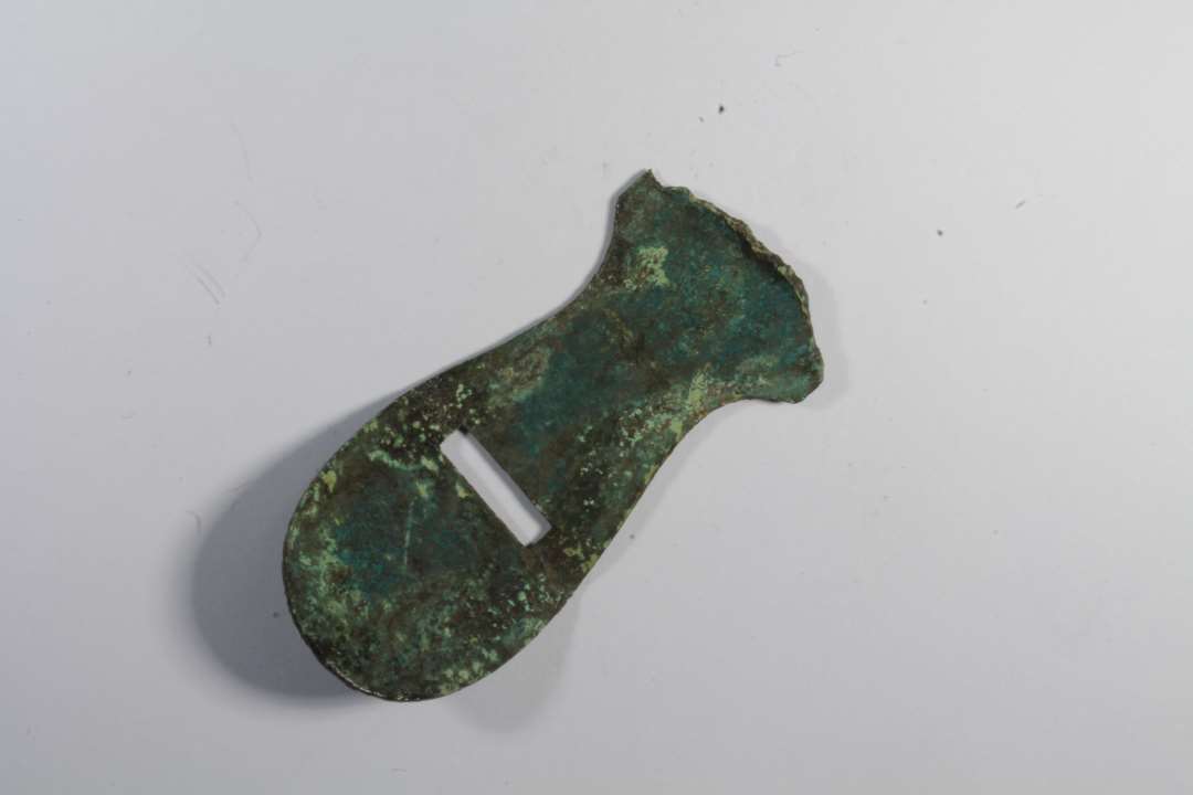 Fragment af bronzegenstand. Flad, aflang, tungeformet med rektangulært hul. Længde: 4,5, bredde: 2,2 cm.