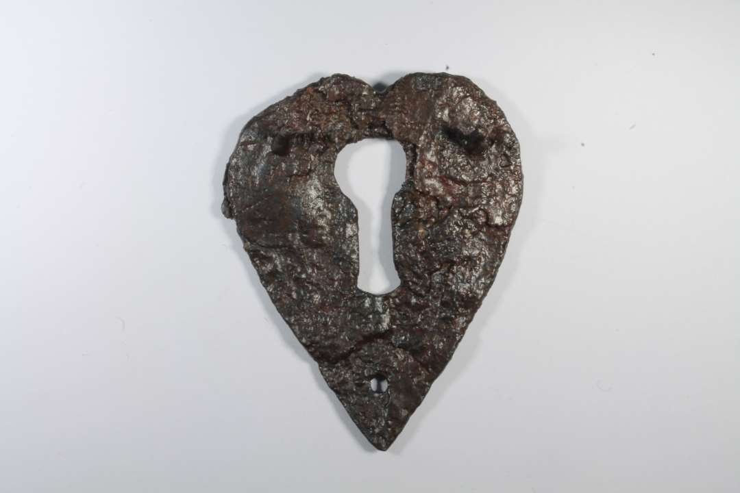 Hjerteformet nøgleskilt af jern. Med to isiddende nitter og et nittehul. Højde: 6,3 cm., bredde: 5,2 cm.