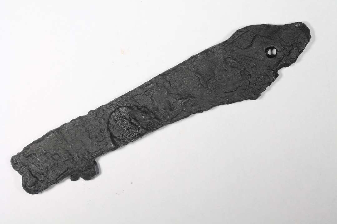 Fragment af flad, aflang jerngenstand, måske noget fra et knivskæfte? Med (nitte?) hul i den ene ende. Længde: 9, største bredde: 2 cm.