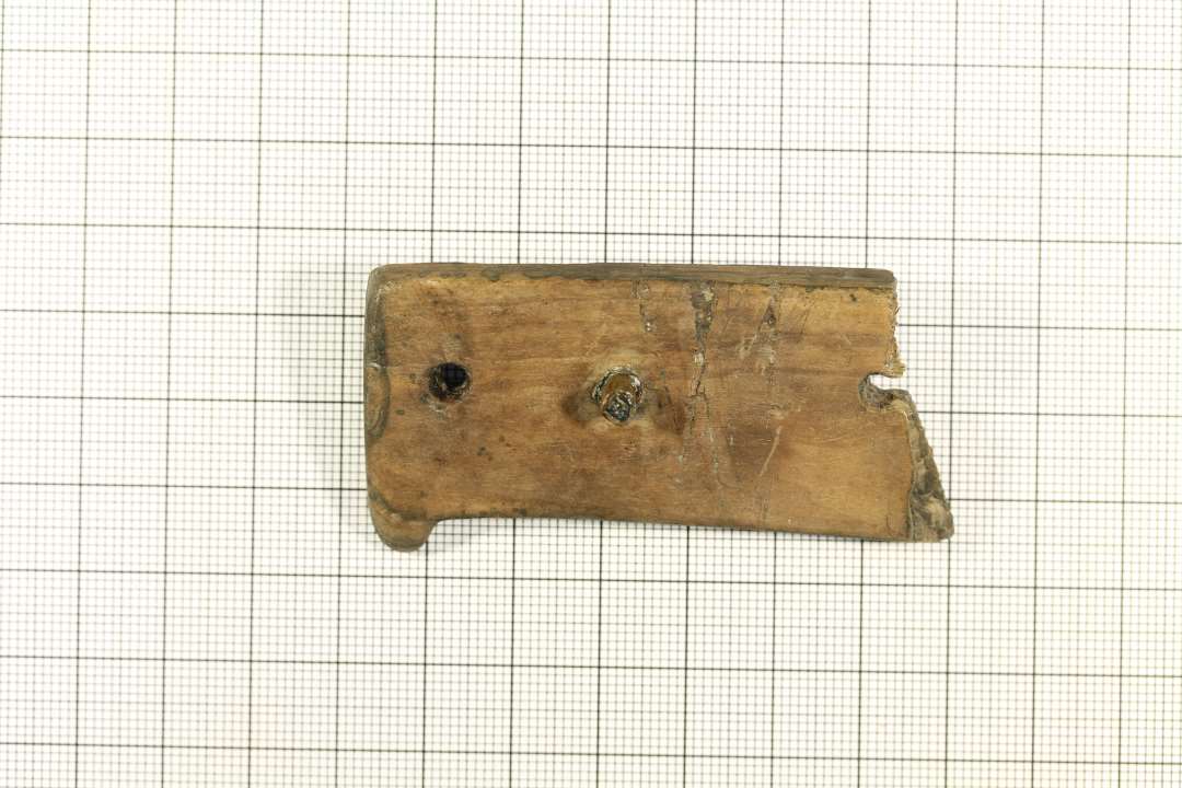 Fragment af trægenstand. Rektangulær form med tungeformet udvidelse i den ene langside, samt isiddende bronzenitte og to tomme nittehuller. Mål: 6,8x3,2 cm.