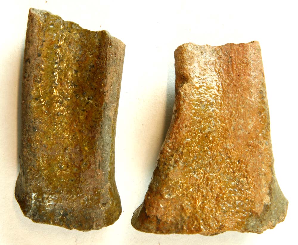 To hankefragmenter af oval tværsnitsform med flad fure på oversiden fra kande af rødbrunligt lergods med gråbrændt kerne. Ydersiden er dækket af ujævn, grønlig blyglasur.