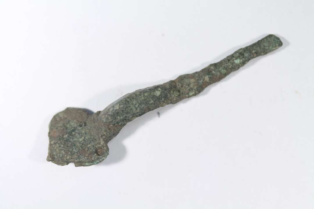 Korsformet fibula af bronze, det er en særfom med gruber i hjørnerne af hovedpladsen, og eventuelle knopper på hovedpladen er ikke bevaret. Ældre Germansk Jernalder. Længde: 7,5 cm., Største bredde: 2,2 cm.