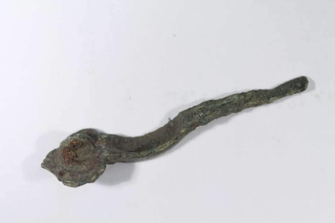Korsformet fibula af bronze, det er en særfom med gruber i hjørnerne af hovedpladsen, og eventuelle knopper på hovedpladen er ikke bevaret. Ældre Germansk Jernalder. Længde: 7,5 cm., Største bredde: 2,2 cm.