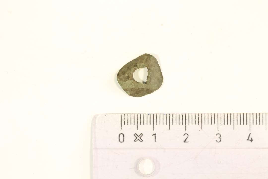 Cirklært stykke bronzeblik med større hul imidten. Lettere krøllet. Diameter: 1,4 cm. Måske fra knivende? 