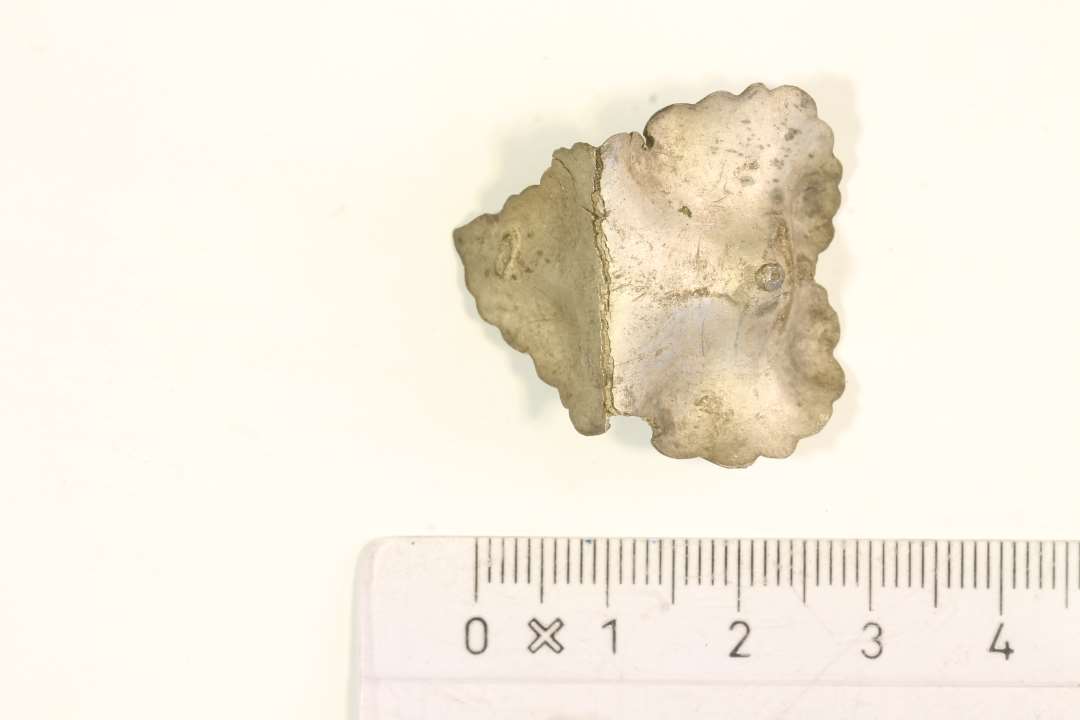 Fragment af broche af metalblik. Udformet som et blad. Rest af nålefæste bagpå. Let sammenkrøllet. Mål: 3x2,8 cm.