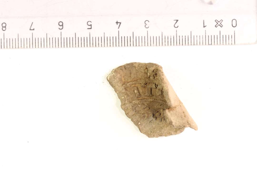 Fragment af stor klædeplombe med omskrift. Største mål: 4 cm.
NM: Fragment af klædeplombe eller emblem af bly med rester af udvisket motiv og omskrift.