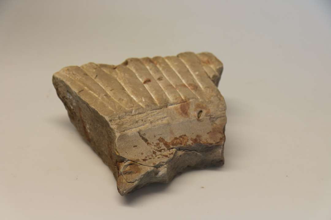 1 fragment af svær kværnsten af grålig sandsten, finkornet med rødlige årer. Største mål : 27 x 23,5 tykkelse : 11 cm. Stenens underside er fladt afbanet, den udadvendende side er prikhugget, mens oversiden har grovhuggede, eadiære stråler samt i midten resten af en skålfomet forbygning. Overflade samt indersiden af fordybningen er glat afslidte. Stenen fandtes sekundært anvendt som del af gulvlaget A5 i huset, jvf. beskrivelser, opmålinger og fotos. Lag A5.