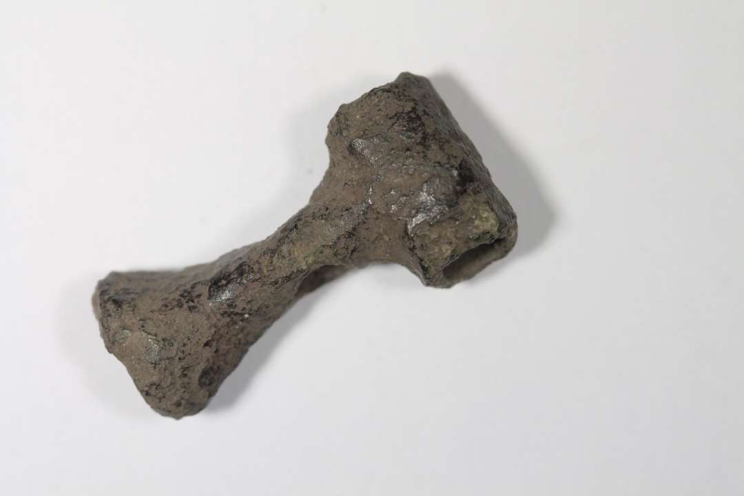 T-formet genstand af bronze med hul i hver ende af/gennem overliggeren og et hul i samme retning gennem stilken. Enden af stilken er muligvis en støbekegle. Længde 2,94 cm. Bredde 1,49 cm. Jernalder eller Middelalder