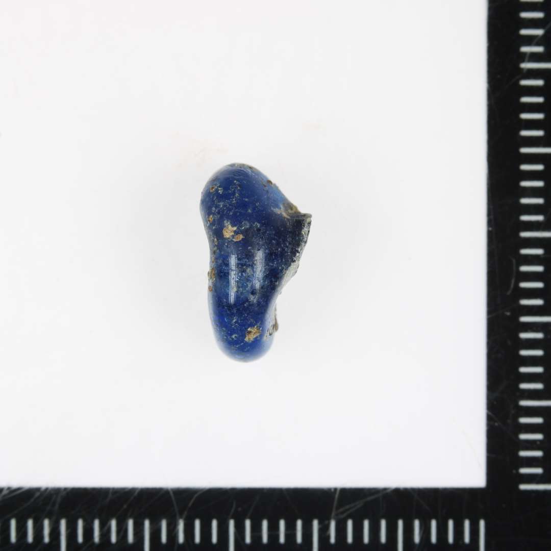 Blå, halv ringformet perle, der har hængt fast på jerndornen og dermed er hullet trukket skævt og formentlig derved er perlen knækket af.