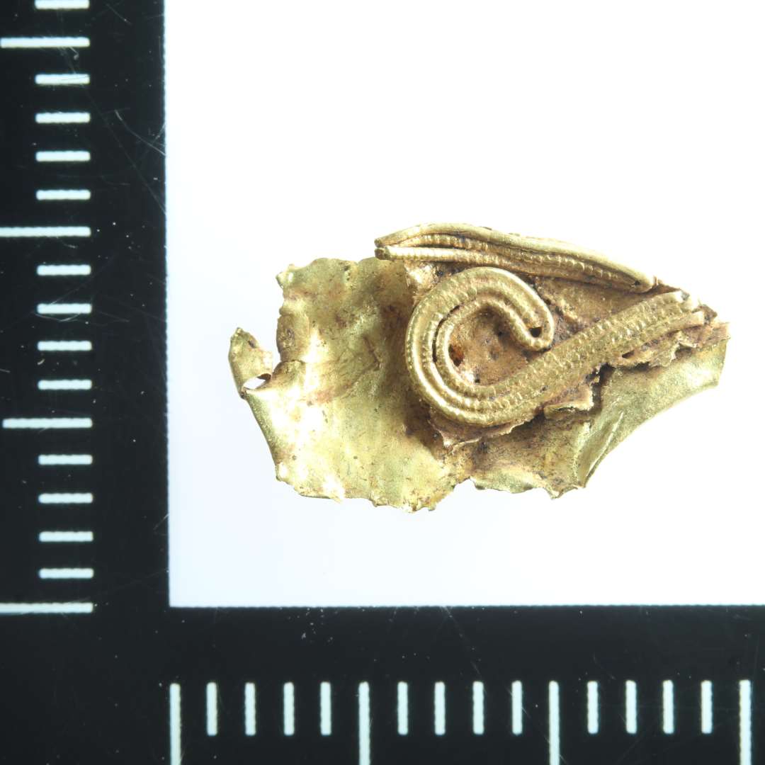 Mindre fragment (1,3 x 0,7 cm) af guldblik ornamenteret med filigran. Længde: 1,2 cm.