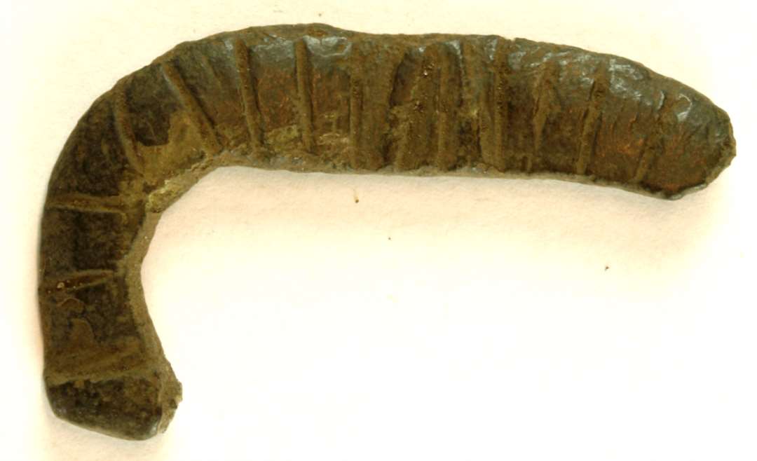 Fragment af bælte- eller skospænde af kobberlegering. En del af rammen bevaret. Dekoreret med stregornamentik. Største mål: 43 mm.