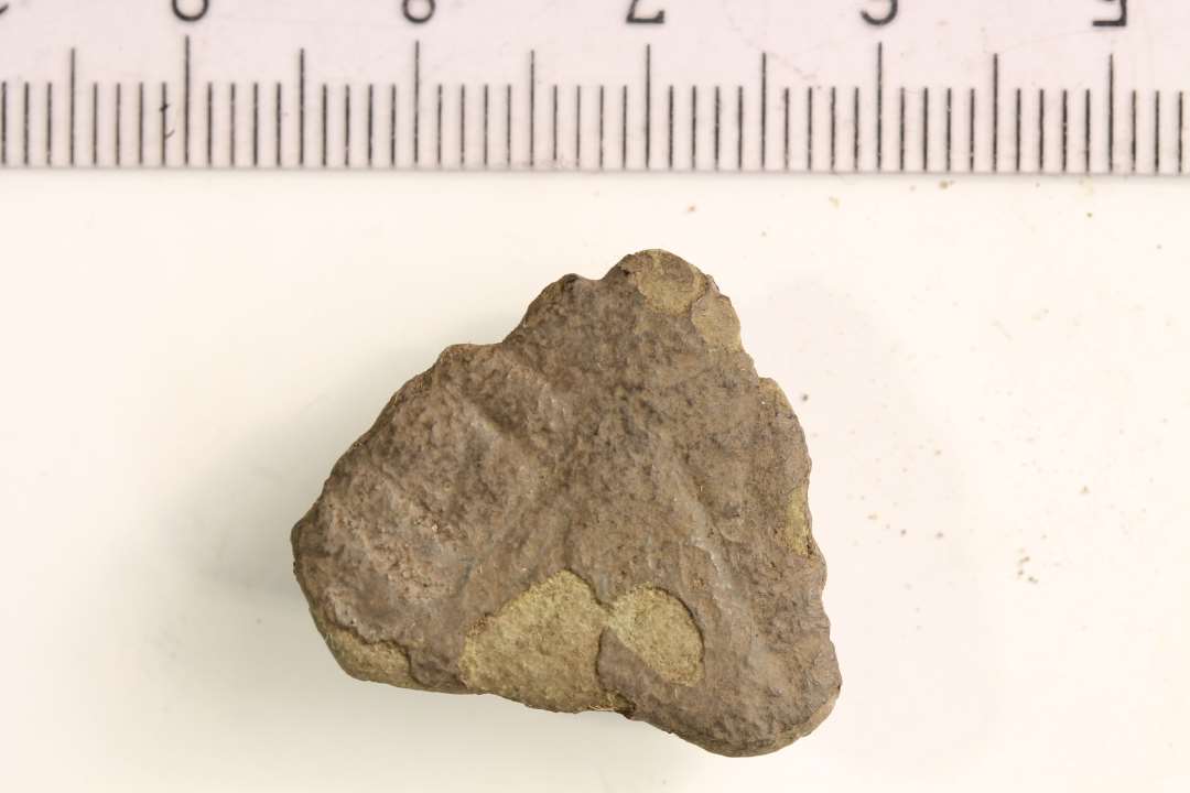 Midterfeltet af en trefliget fibula med slidt overflade, der kan ses rester af geometrisk stregornamentik. På bagsiden er bevaret nålefæste med rest af jern fra nålen. Største mål: 2,5 cm 