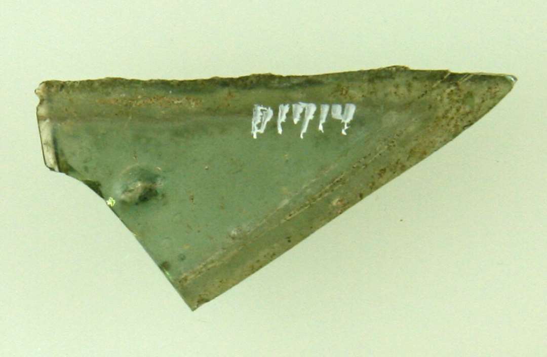 Fragment af spidsen af rude antagelig i form af ligebenet trekant af grønligt glas, den ene kant er glat tilskåret, den anden er afnappet. Største længde: 3,75 cm. Højde: 1,8 cm.