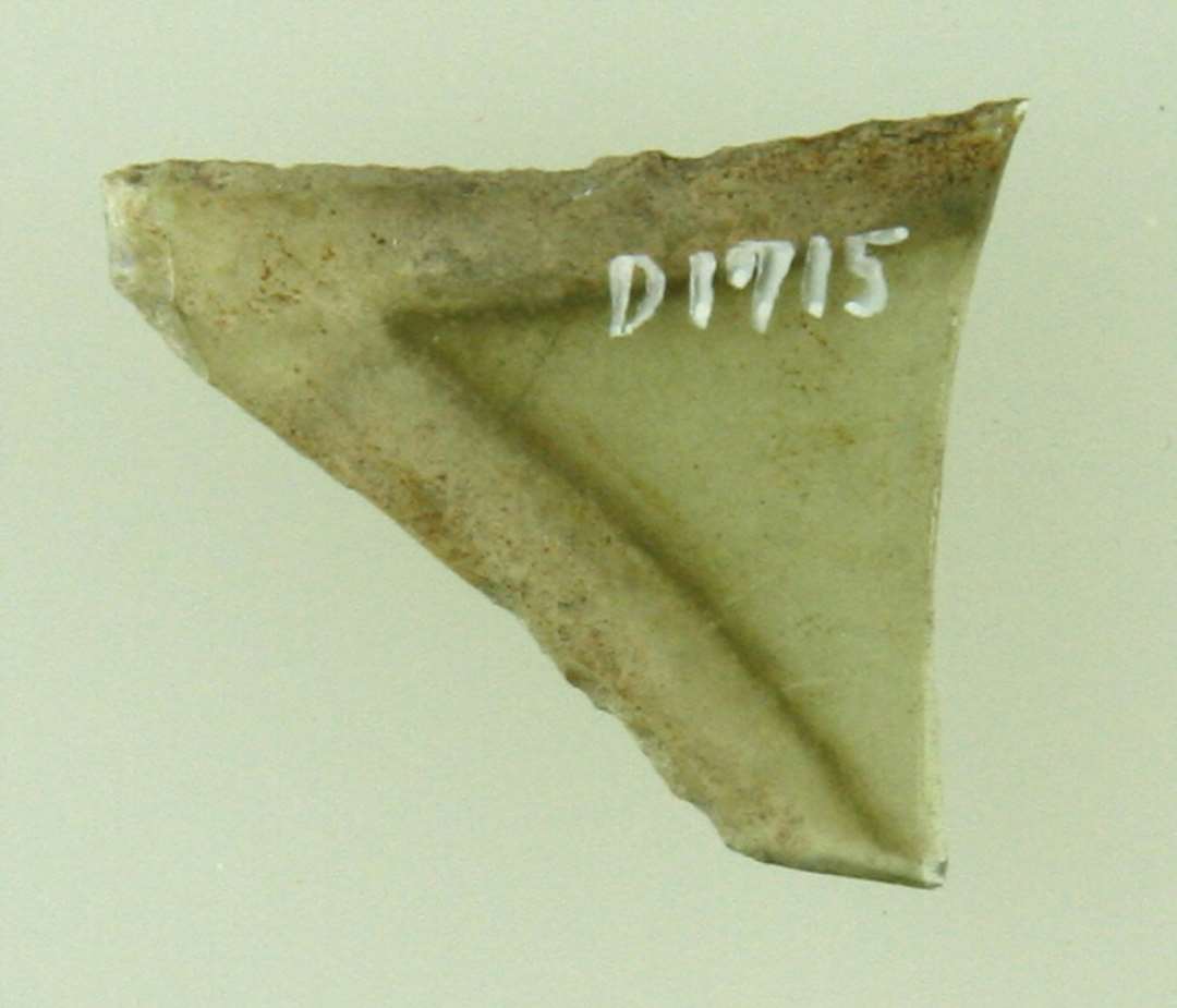 Fragment af spidsen af rude af grønligt glas med let konkave afnappede kanter.  Længde: 2,55 cm. Bredde: 2,2 cm.