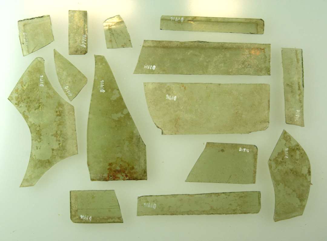 14 fragmenter af ruder af klart grønligt glas, alle med et retvinklet hjørne bevaret, 8 af fragmenterne viser spor af glat tilskårne kanter, 4 af afnappede kanter. Største mål: 2.5-10 cm.