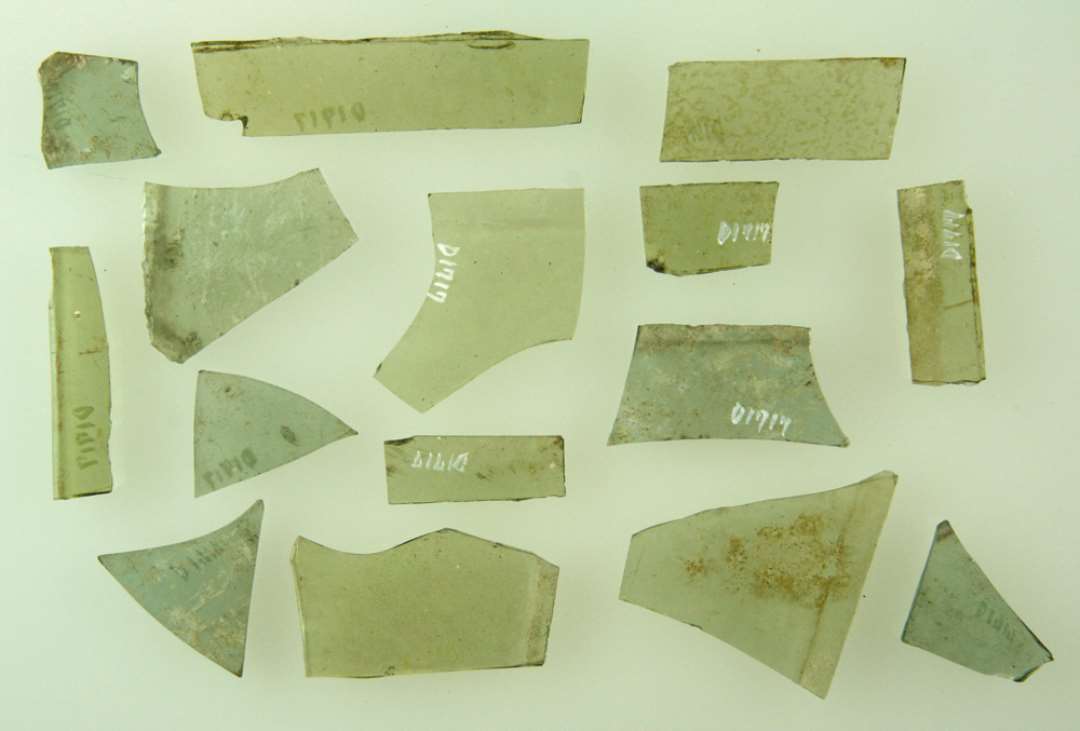 17 fragmenter af ruder af grønligt glas af ubestemmelig form heraf 15 med spor af glat tilskårne kanter, 2 med spor af afnappede kanter. Største mål: 1,7-6,2 cm.