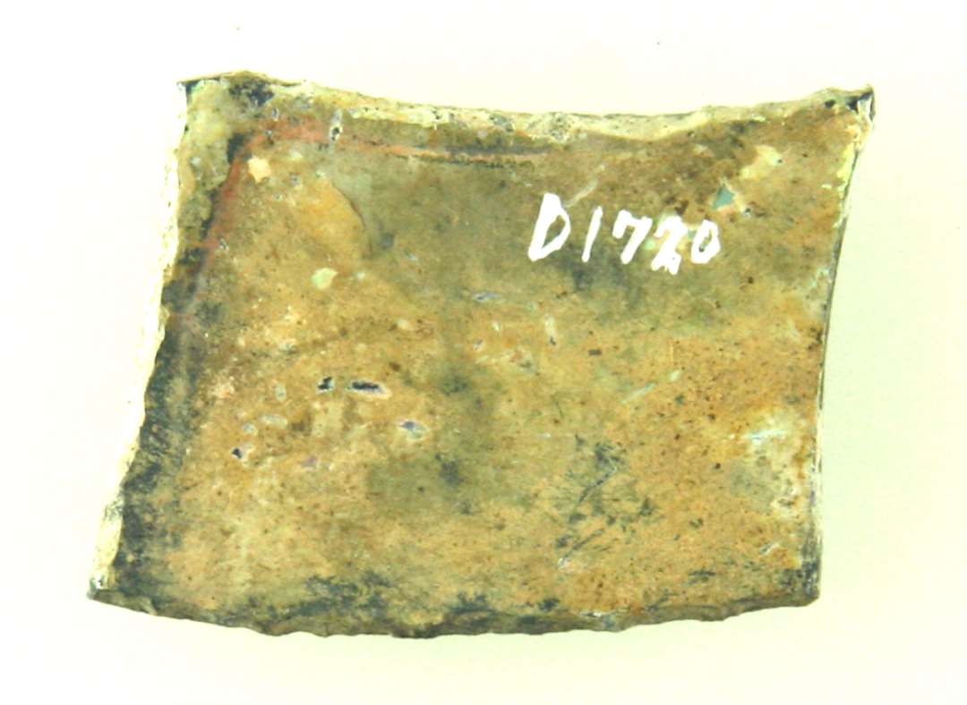 Fragment af båndformet rude af blåligt glas med påmalet rankemønster i brun farve. Ruden har haft trapezagtig form med to krumme langsider. Største mål: 3,2 cm.