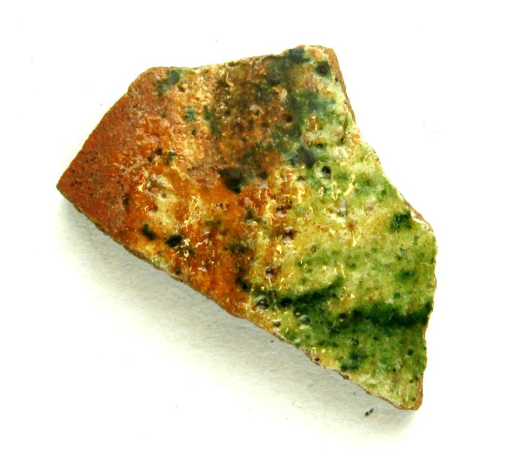 Bugskår af rødbrændt, flamsk lergods, der på ydersiden er delvis dækket af pibelersbegitning, hvorover spættet grønligt blyglasur.