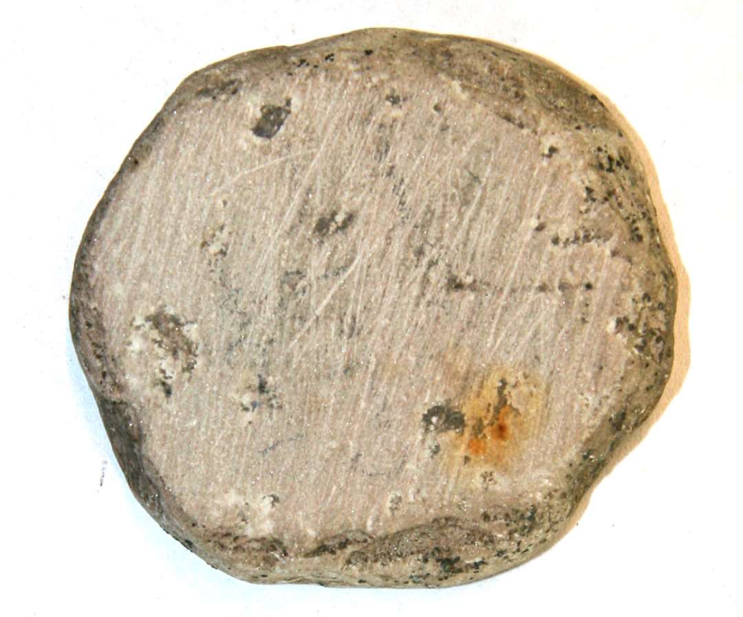 En skive grå sandsten. uregelmæssig, tildannet cirkulær form. Mål: Diameter 5 cm., tykkelse 0,8 cm. Slibestriber på den ene side.