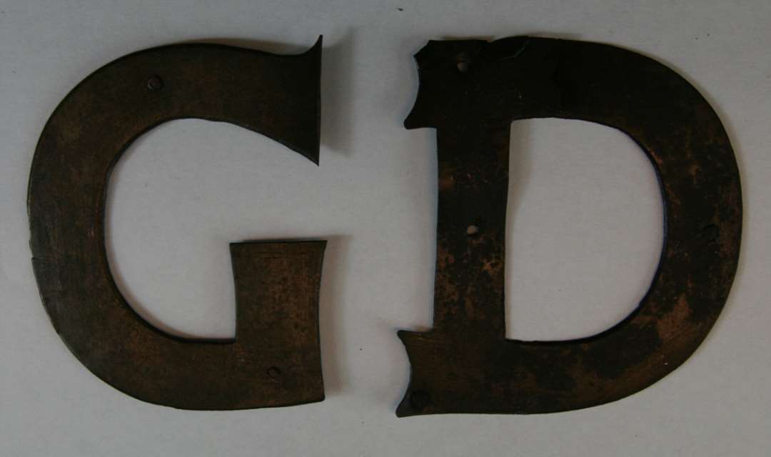 2 separate bogstaver ( G og D ) i form af store latinske bogstaver, udført i nu irret bronzeplade. Bogstaverne har været fastholdt med bronzestifter. Mål : 7,8 x 6,4 cm ( G ) - 8 x 7,3  ( D ).