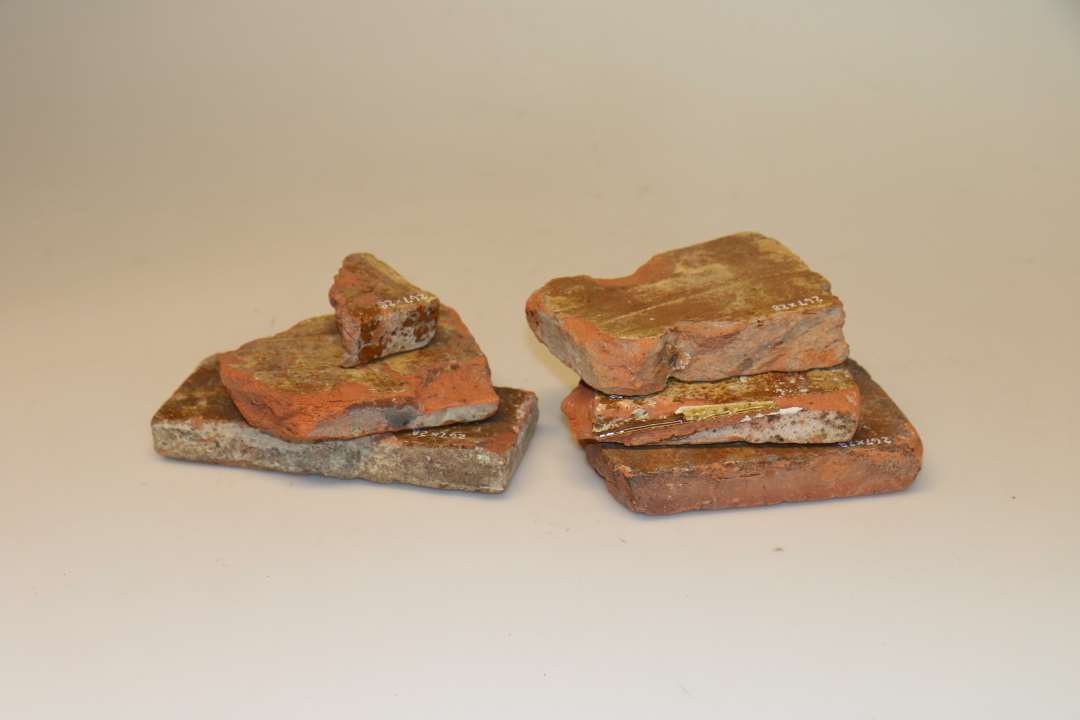 Flisefragmenter, ler. 6 fragmenter af flisetype af rødbrændt ler, hvis overside er dækket af uregelmæssigt påstrøget og kun delvis dækkende pibelersbegitning, hvorover igen klar blyglasur. Eneste oprindelige sidemål : 18,7 cm. Tykkelse : 2,7 cm.