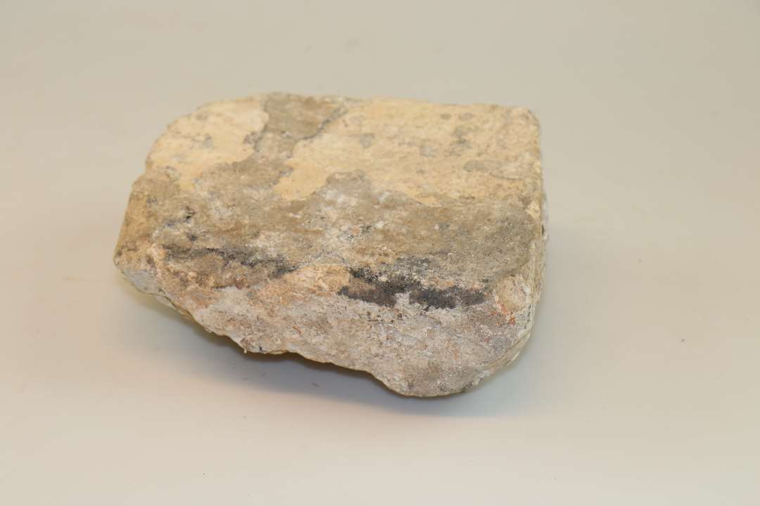 Bygningssten. En sten af ret skæv rektangulær form af hvidgrå kalkagtig stenart, planhugget på den ene fladeside med tilhugne sidekanter og ujævn utildannet bagside. Sidemål: 19 x 21 cm. Tykkelse : ca. 9 cm.