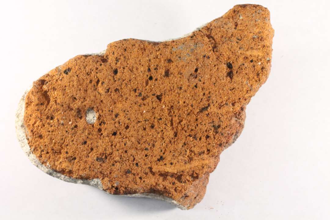 Bygningssten. Et fragment af rødbrændt ler. Den bevarede ende af stenen  har omtrent halvcirkulær form, der via en lille afsats fortsætter i indsvungne sider, der igen går over i en hvælvet form i stykkets ene side, hvor der findes spor af en lille fals. Største bevarede længde : 13 cm. Tykkelse : 8,5 cm.