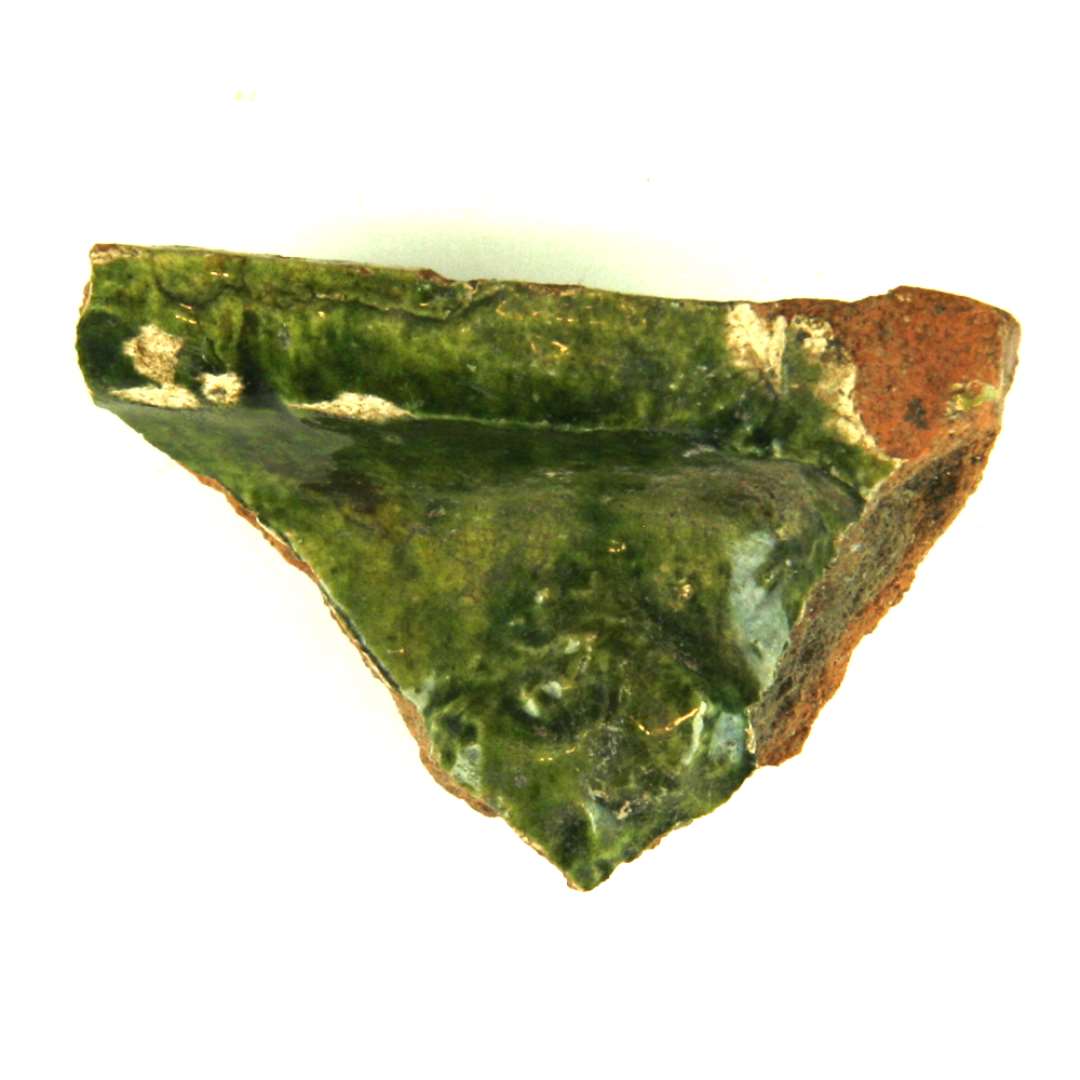 Fragment af kakkel, grønglaseret, hjørnestykke. I midten rest af relief (mandshoved), ramme med søjleværk. Største mål: 6,3 cm.