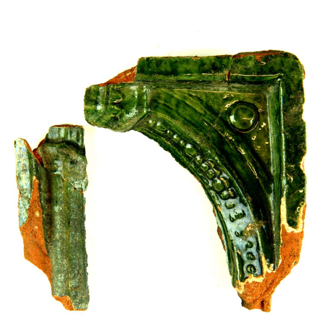 To fragmenter af grønglaseret kakkel hver sammenlimet af flere stykker. Øverste højre hjørne. Søjlestilling med bueslag, hvorpå perlestav. I svinklen halvkugle. Høj yderramme. 