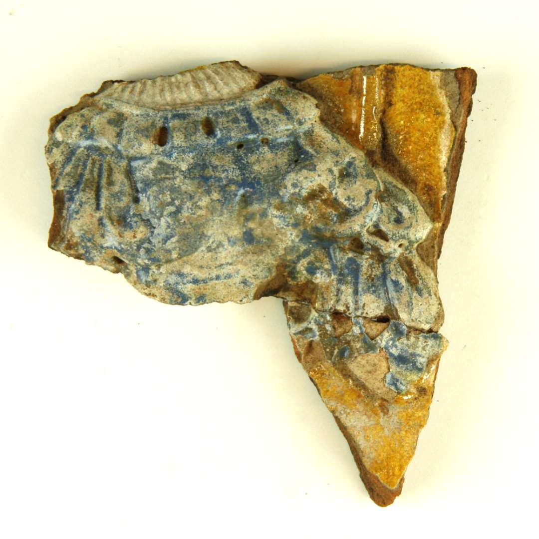 To sammenlimede fragment af polykrom, lodret kakkel. Midterstykke, samlet af 2 stykker. Brystpartiet og venstre ærme af kvindefigur i blå kjole, med hvidt lin i halsen. Største mål: 9,5 cm.