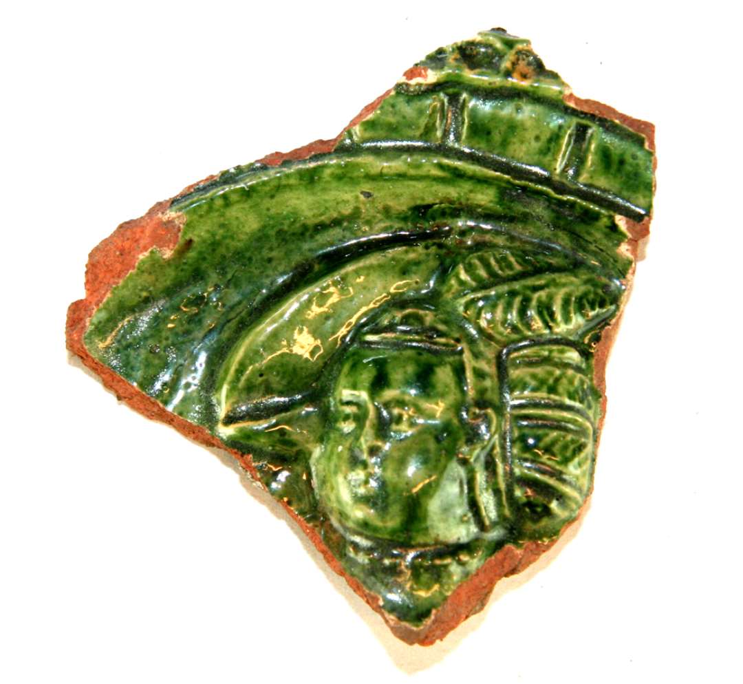 Stykke af grønglaseret kakkel. Største krydsmål: 7,2 x 7,4 cm. Glasuren lyst grøn, godset rødt med sværtning på bagsiden. På fragmentet et motiv med et kvindehovede med kunstfærdig frisure bag venstre øre og baret med fjer over højre. Et højtsiddende halslin ses.