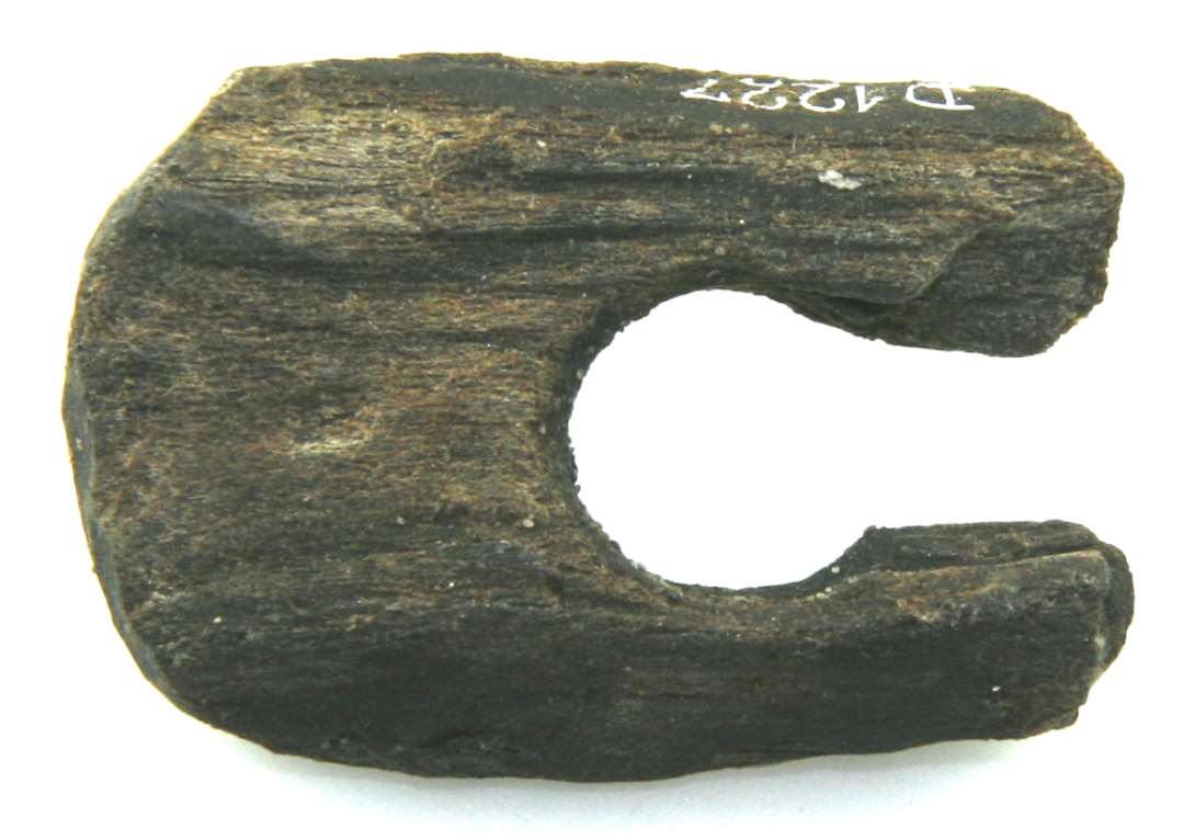 Fragment af fladt tildannet træstykke afrundet i begge ender og forsynet med en gennembroing på ca. 1,5 cm. i diameter på midten. Et parti mellem midterhullet og stykkets ene ende er afbrudt.  Største længde: 5,1 cm. Største bredde: 3,5 cm. Største tykkelse: 1 cm.