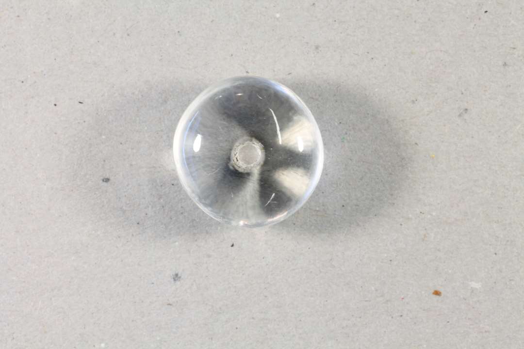 Perle af bjergkrystal. Form som en fladtrykt kugle, tykkelse 1,3 cm. Diameter 1,7 cm. Forsynet med let konisk gennemboring med små af sprængninger i hullets smalleste ende. Perlen er forsynet med 3 omløbende riller.