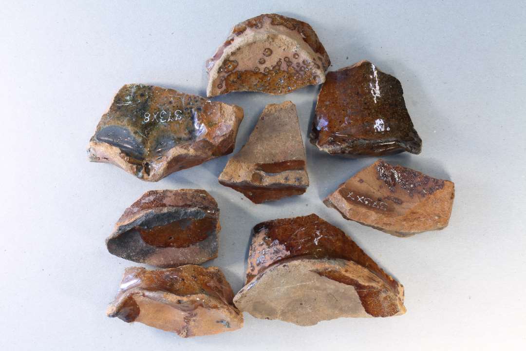 8 bundskår af rødbrændte, klart blyglaserede kander med større og mindre hvide sten i magringen. Svarer til det lertøj, som formodes at være lavet lokalt, antagelig efter ca. 1300. B1.