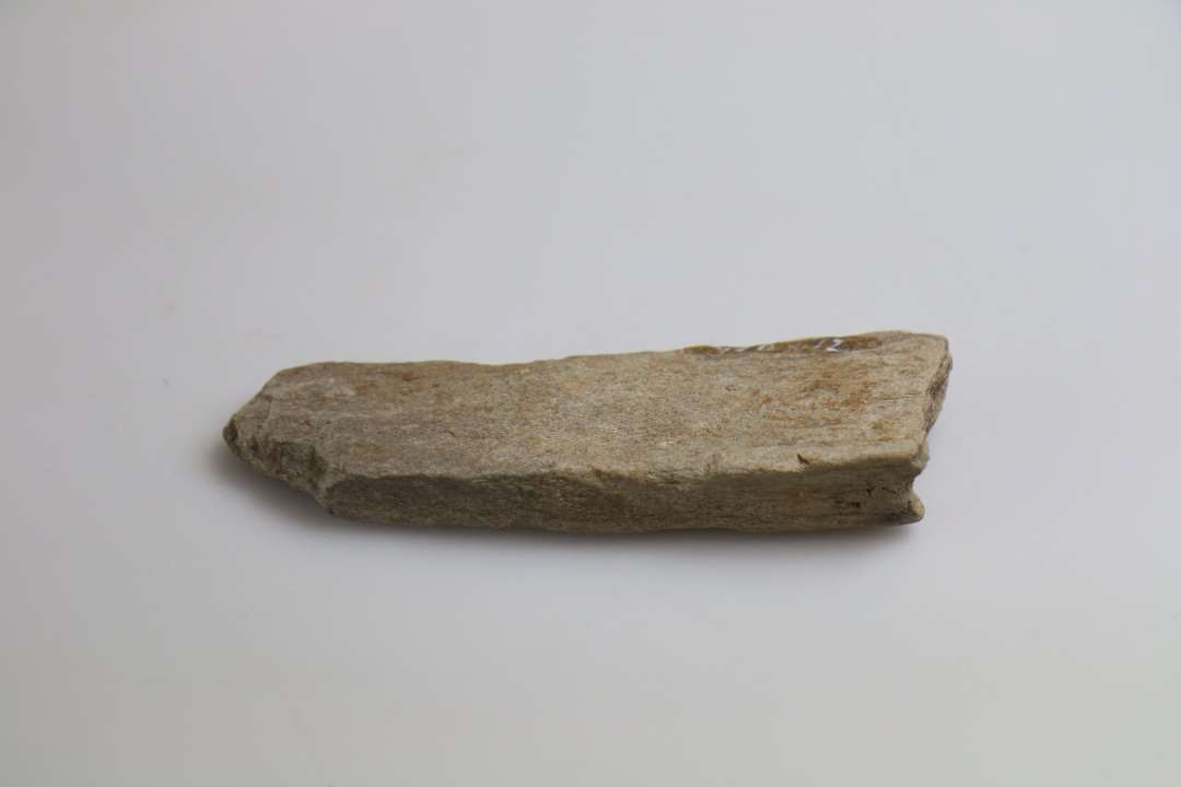 1 fragment af hvæssesten af gråligt, finkornet sandsten, uden synlige slidespor, afbrudt i begge ender, og smalnende til mod den ene, tilspidsende ende. Længde: 12,3 cm. Største bredde: 3,5 cm. Tykkelse: 1,3 cm.