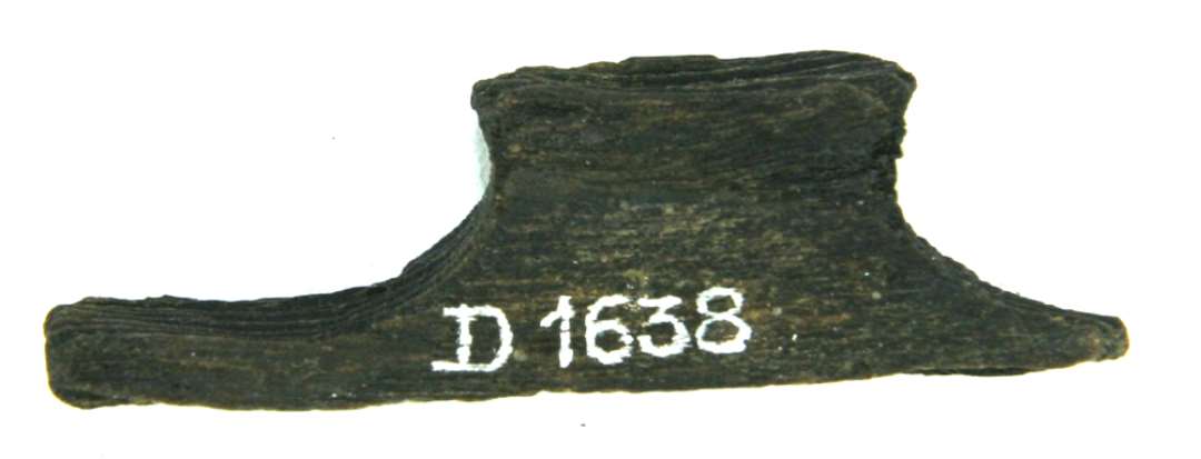 Fragmenteret træstump. Med spor i kanten af to gennemboringer, oprindelig ca. 2 cm. i diameter anbragt med ca. 1,7 cm's mellemrum.  Stykkets længde: 4,5 cm. Bredde: 1,2 cm. Tykkelse: 1,3 cm.