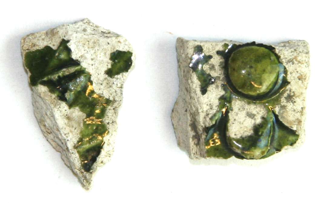 To sideskår af hvidgrå hårdtbrændt pibelersmasse med rester af pålagte bladskælornamenter på ydersiden, der ligesom indersiden er dækket af stærkt afskallet grønlig blyglasur.