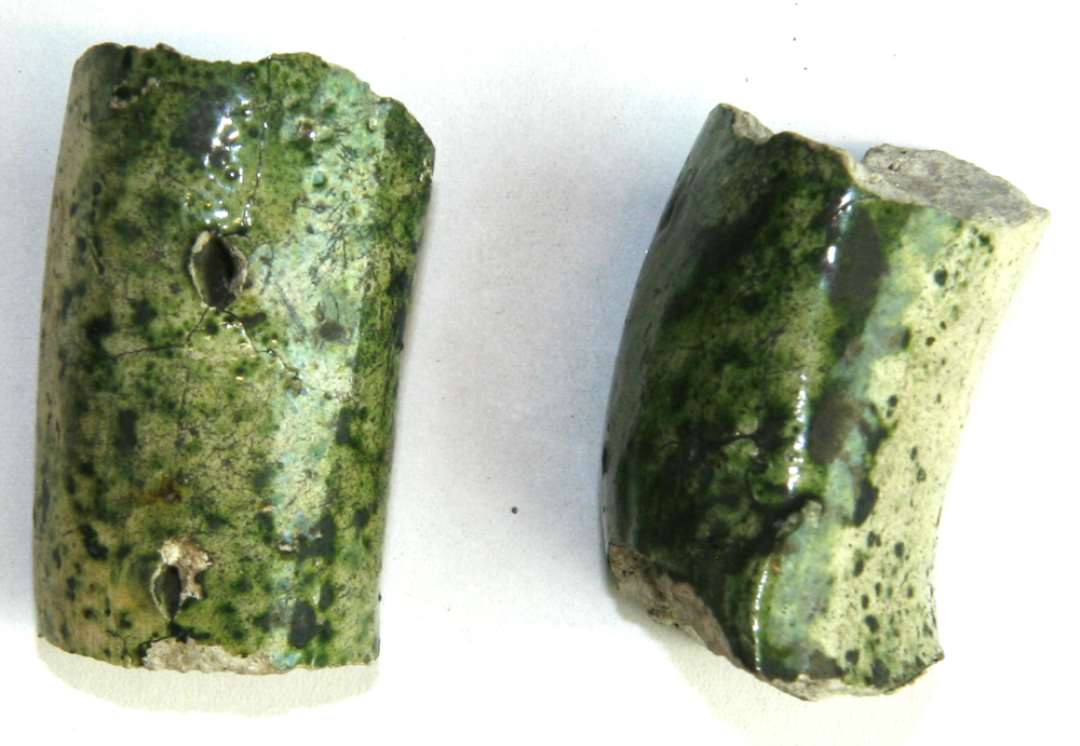 To hankefragmenter af trind pølseform med 2 indstukne huller på oversiden; gråbrændt pibelersmasse med grønspættet blyglasur, der udfylder hullerne på det ene fragment.