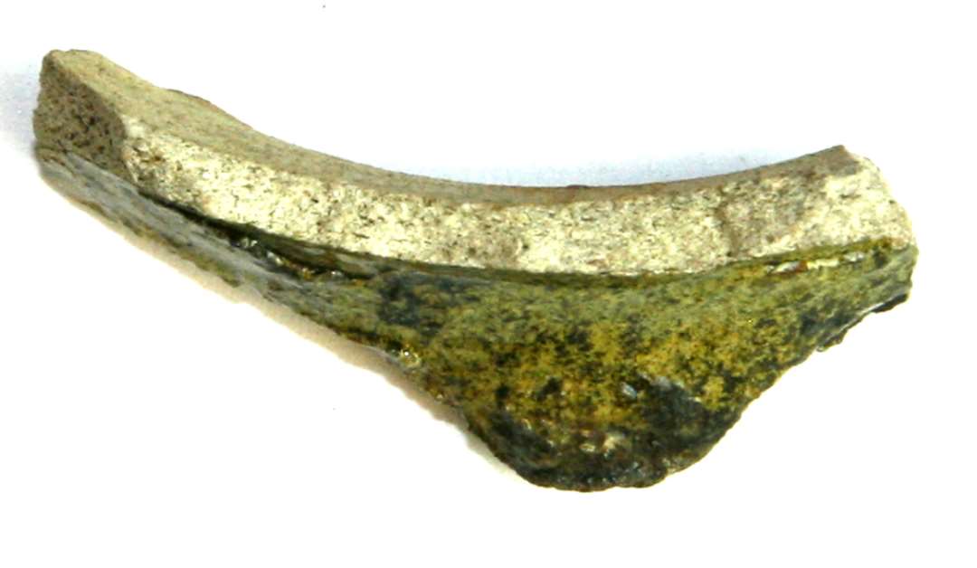 Bundskår af gråbrændt pibeler med grønlig blyglasur på ydersiden. På overgangen mellem den vandrette bundflade og den lodrette side findes en udstående, knoplignende svagt nedadbøjet 'tå'. 