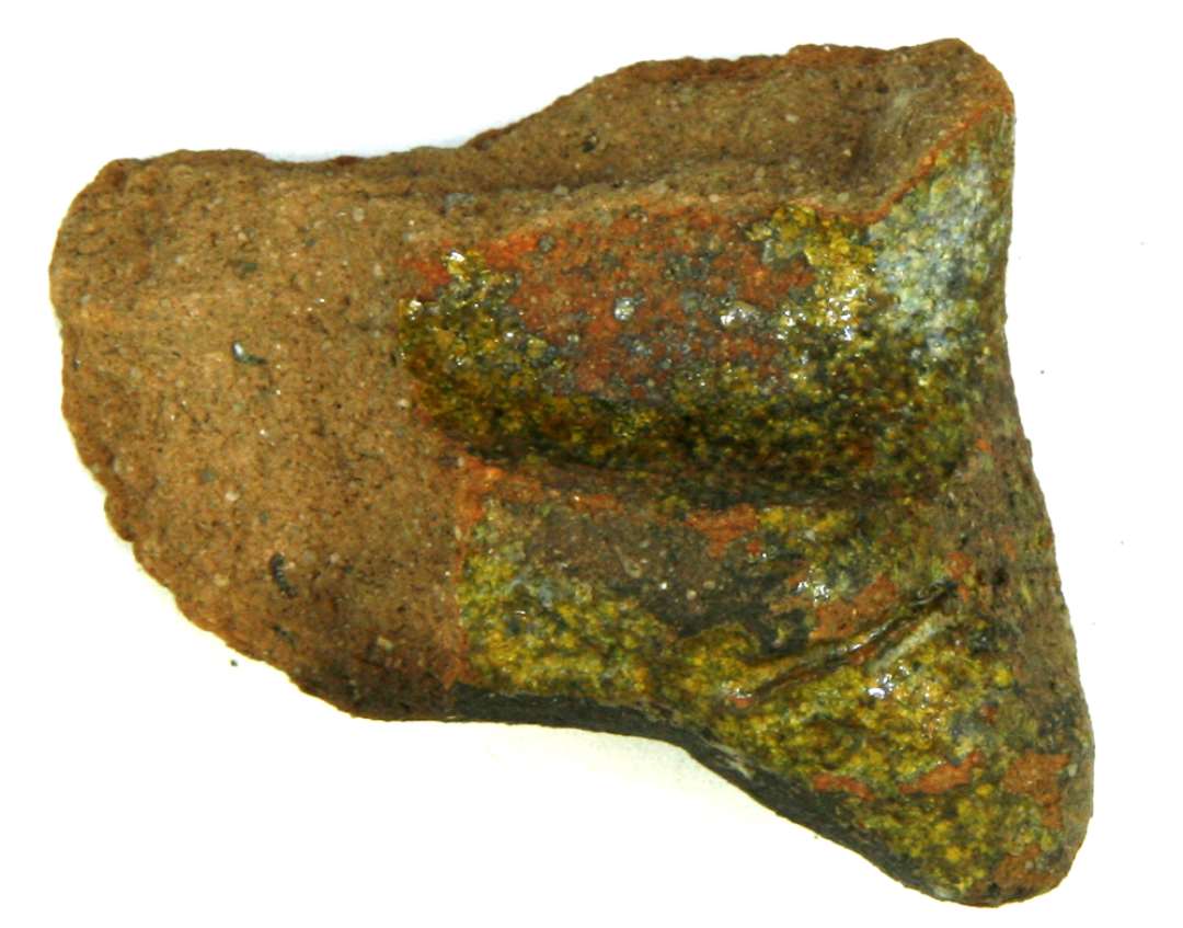 Fragment af gråbrændt lermasse. Muligvis del af hank eller afhældningsrende fra stegefad med knopformet vulst ved den ene side samt rest af dyb hulkehl på midtpartiet. Grønlig plettet blyglasur på oversiden.