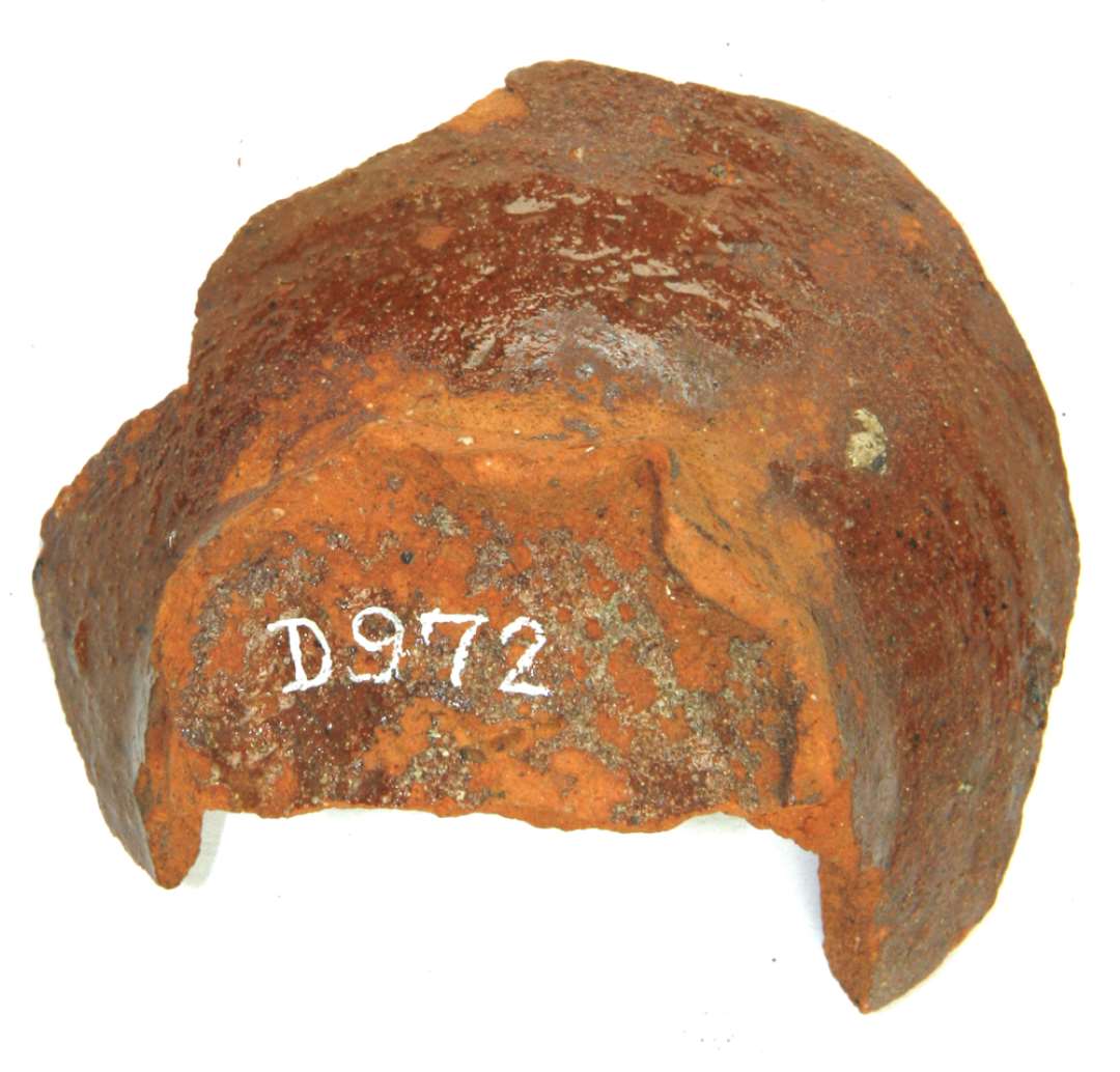 Fragment af underdel af lerkar af rødbrændt lergods med fremstående bølget fodrand, hvorfra karvæggen buer kraftig ud. Ydersiden dækket af klar blyglasur.