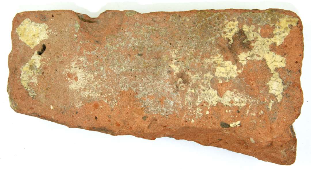 Flisefragment af rødbrændt ler, på oversiden forsynet med stærkt udslidt pibelersbegitning, hvorover klar blyglasur. Fragmentets største længde: 18,3 cm. Ty.: ca 3,2 cm.